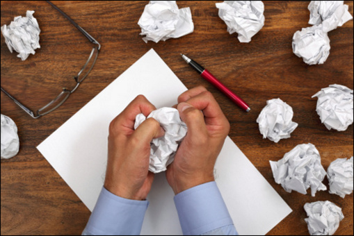 A writer holds a crumpled piece of paper, with more crumpled paper on the desk