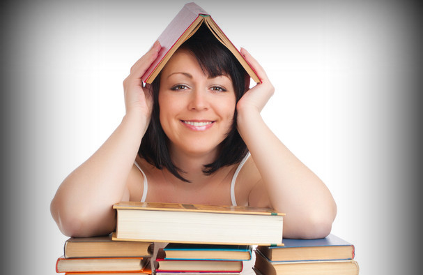 A woman sits behind a stack of books, holding an open book over her head