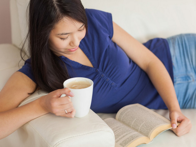 A woman sits on a couch, leaning on the arm, reading a book and drinking coffee.